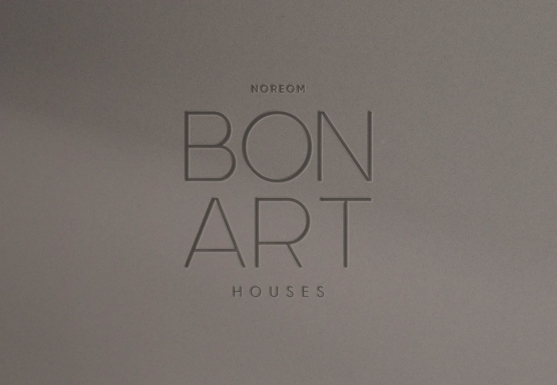 Logótipo gravado em relevo sobre superfície cinzenta com o texto "NOREOM BON ART HOUSES"