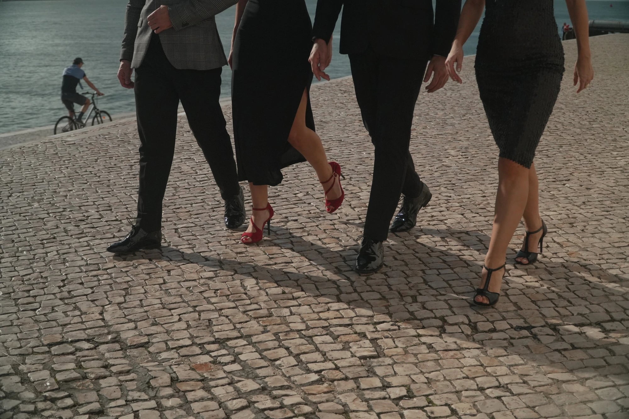 Pernas de quatro pessoas de preto a caminhar em calçada junto ao rio, com saltos vermelhos e sapatos de dança