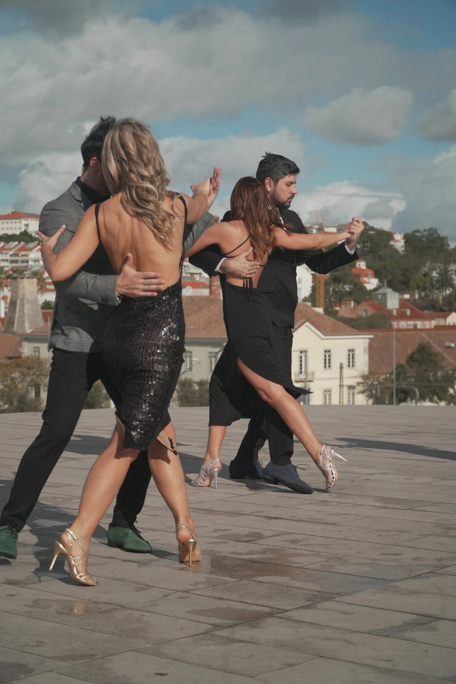 Dois casais vestidos de preto dançam tango numa esplanada de pedra, com telhados de uma cidade ao fundo