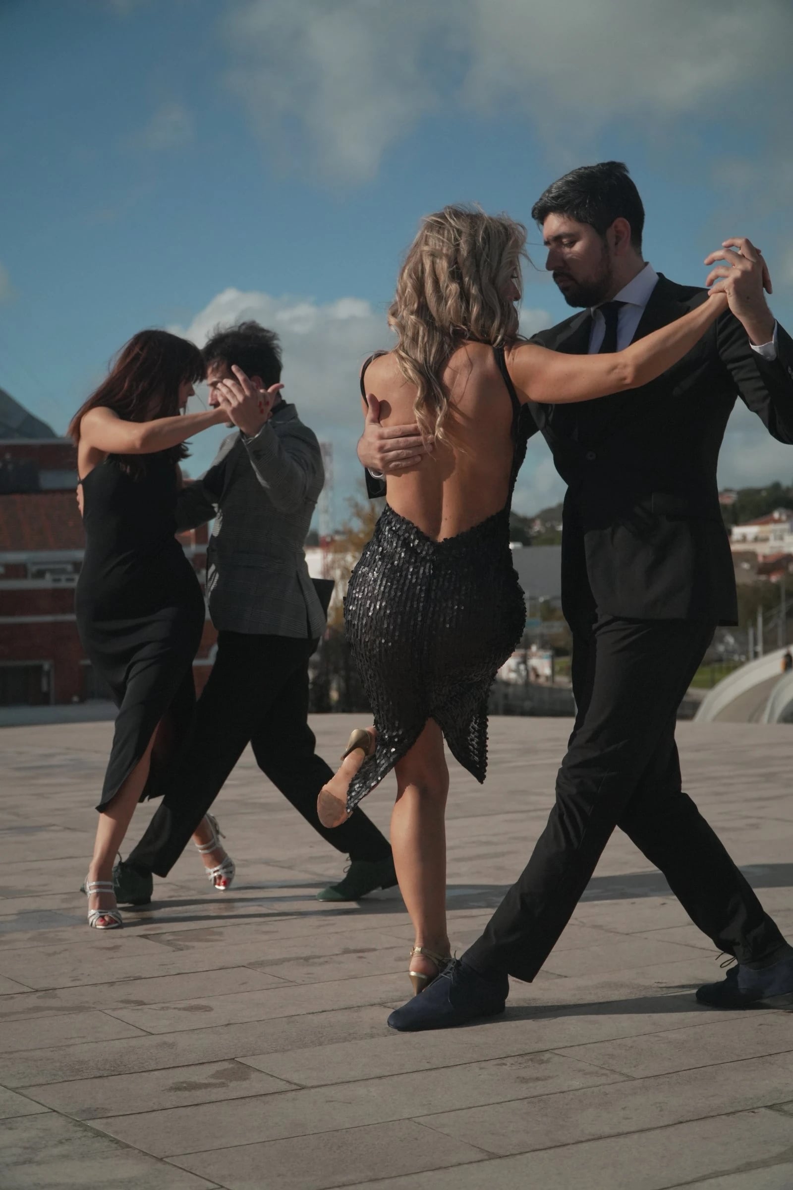 Dois casais em roupa escura dançam tango numa praça pavimentada, sob céu azul com nuvens