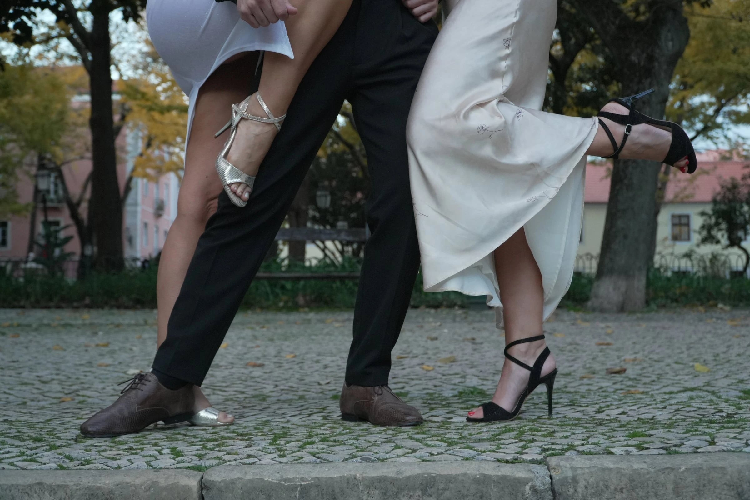 Pernas de três pessoas a dançar tango sobre calçada, com sandálias de salto e sapatos castanhos