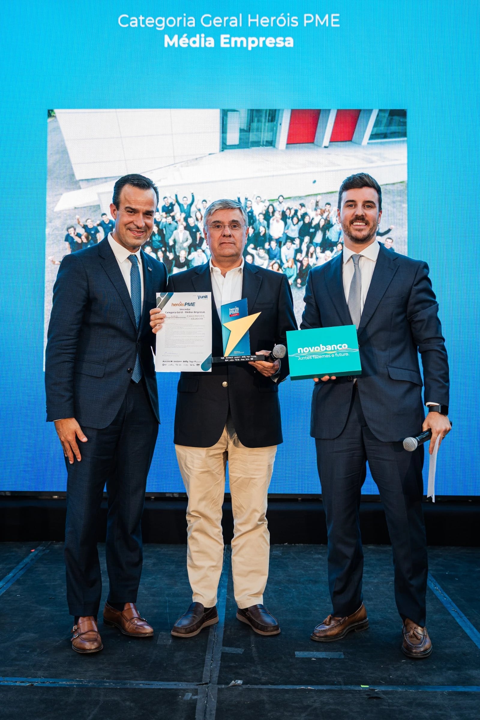 Três homens de fato em palco com diploma e troféu, ecrã atrás com "Categoria Geral Heróis PME – Média Empresa"