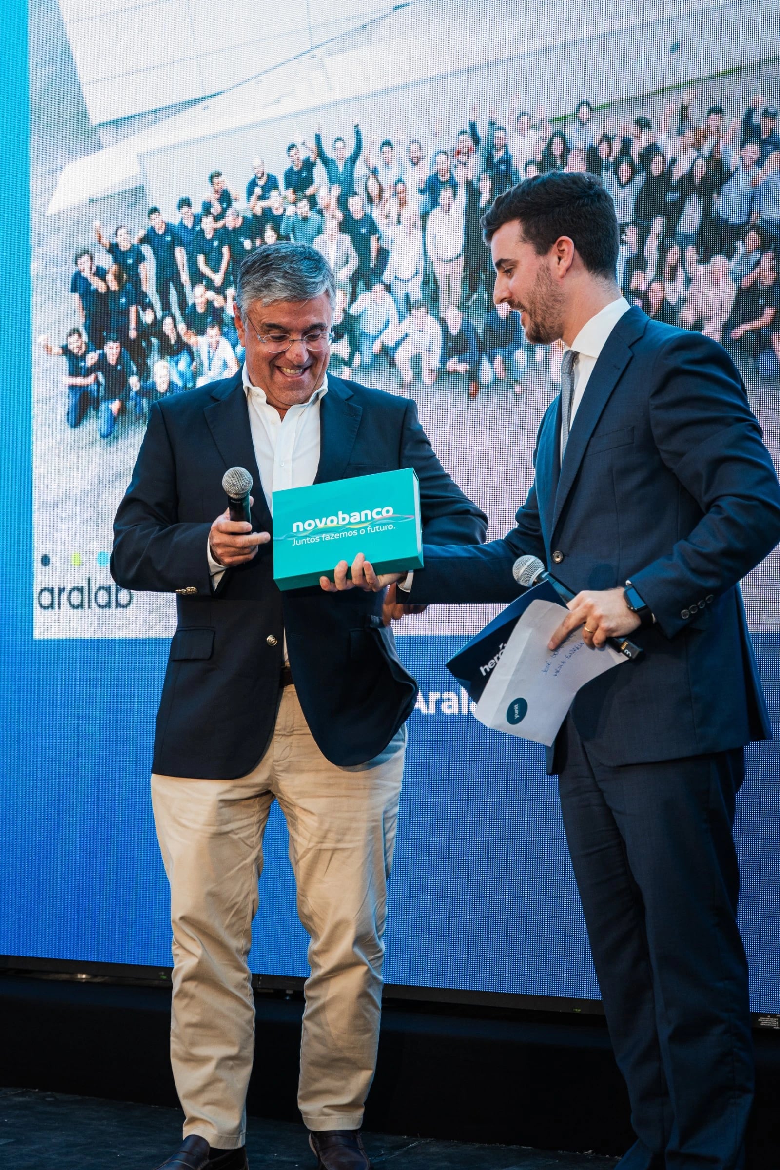 Dois homens de fato em palco entregam uma caixa azul com "novobanco — Juntos fazemos o futuro."