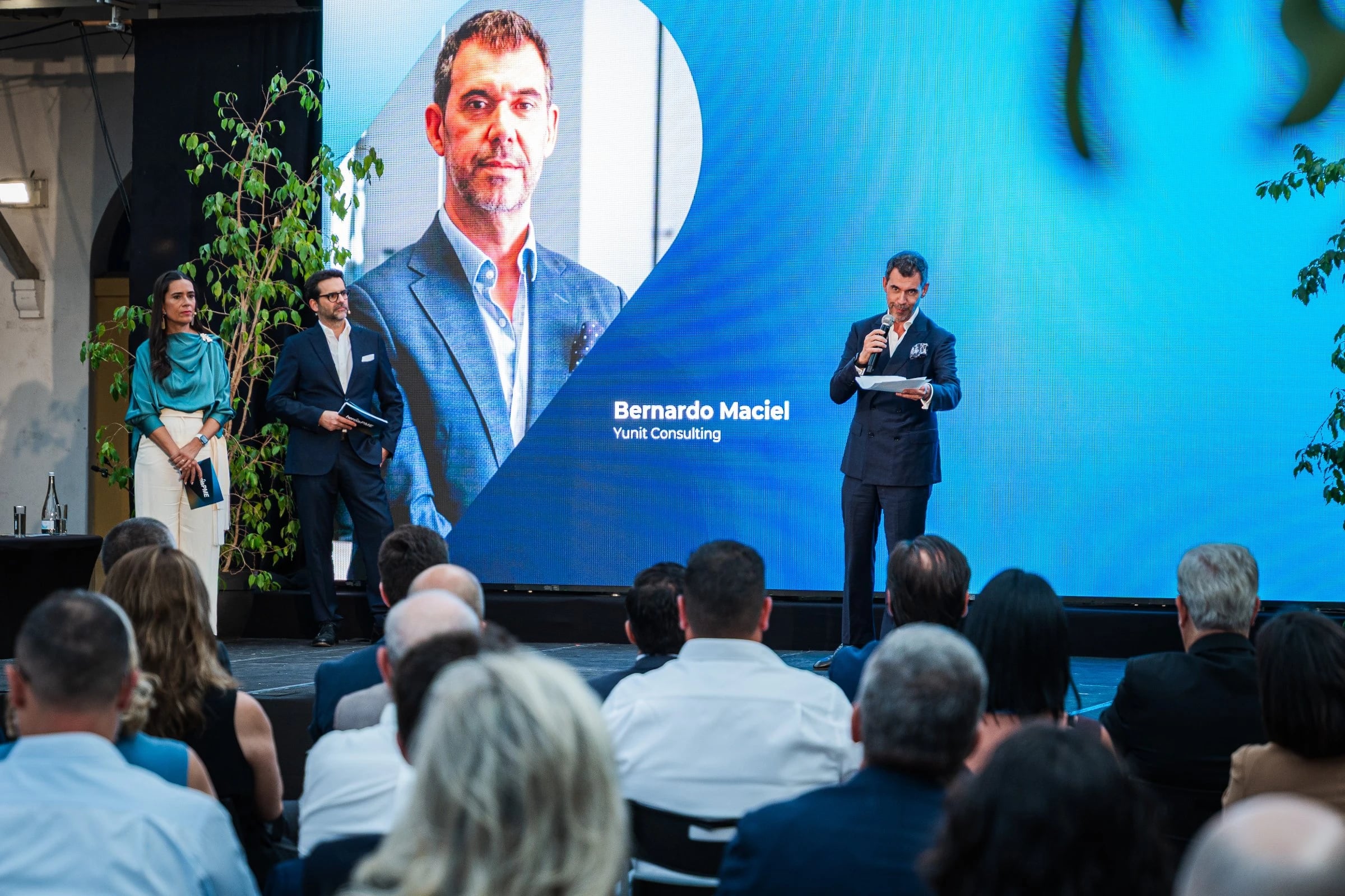 Orador com microfone em palco frente a ecrã azul com foto e o texto "Bernardo Maciel, Yunit Consulting", perante plateia sentada