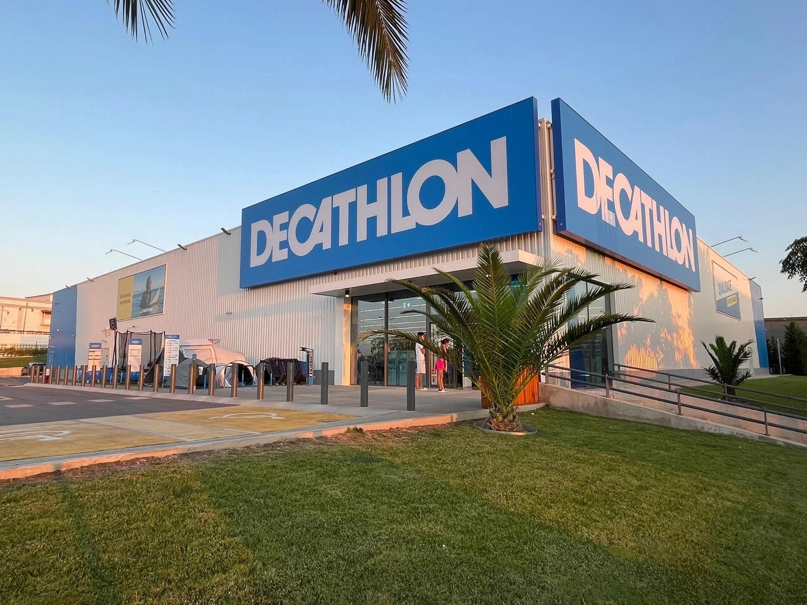 Exterior de uma loja Decathlon ao entardecer, com letreiros azuis "DECATHLON", palmeira e relvado em primeiro plano