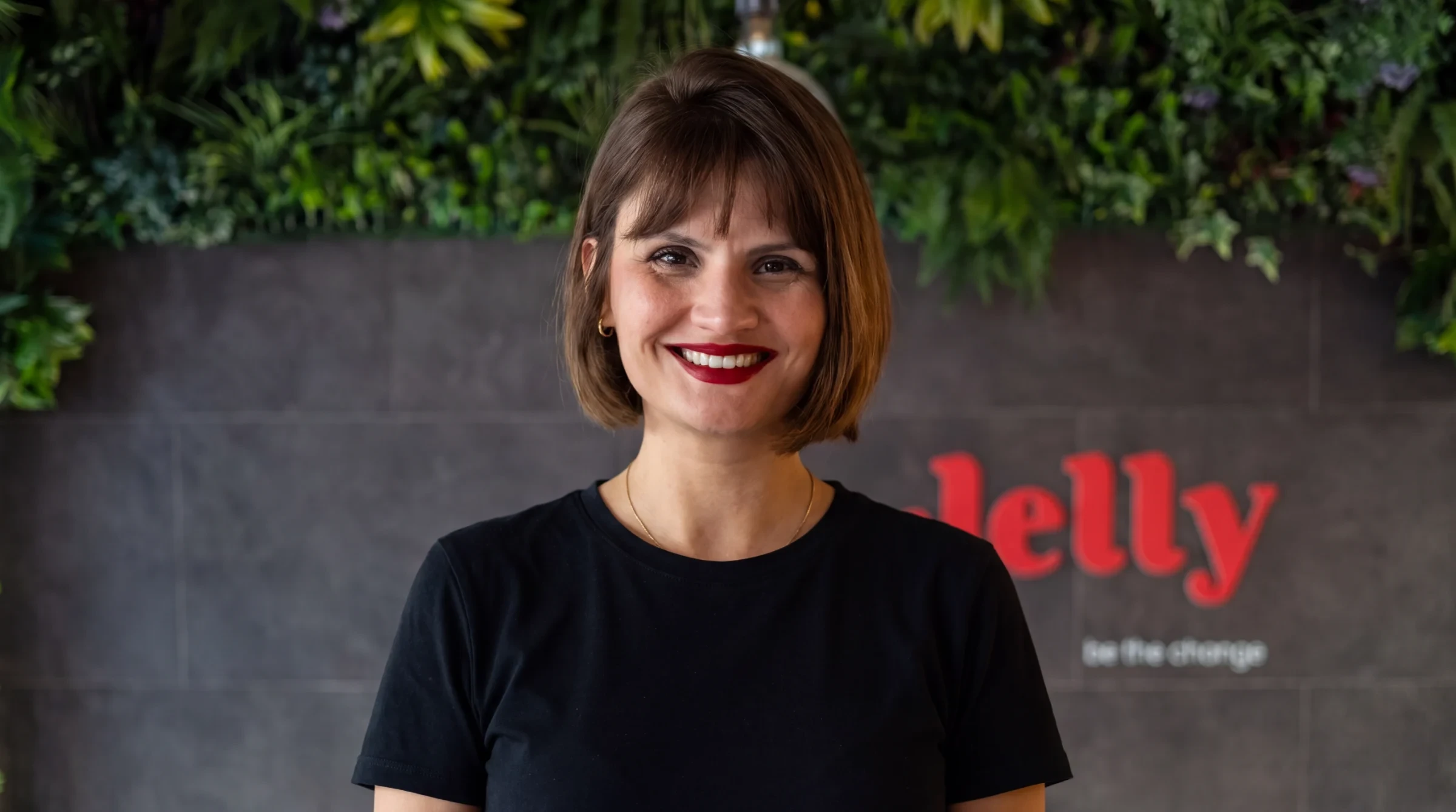 Mulher de cabelo curto castanho e t-shirt preta sorri diante de parede com o logótipo "Jelly — be the change"