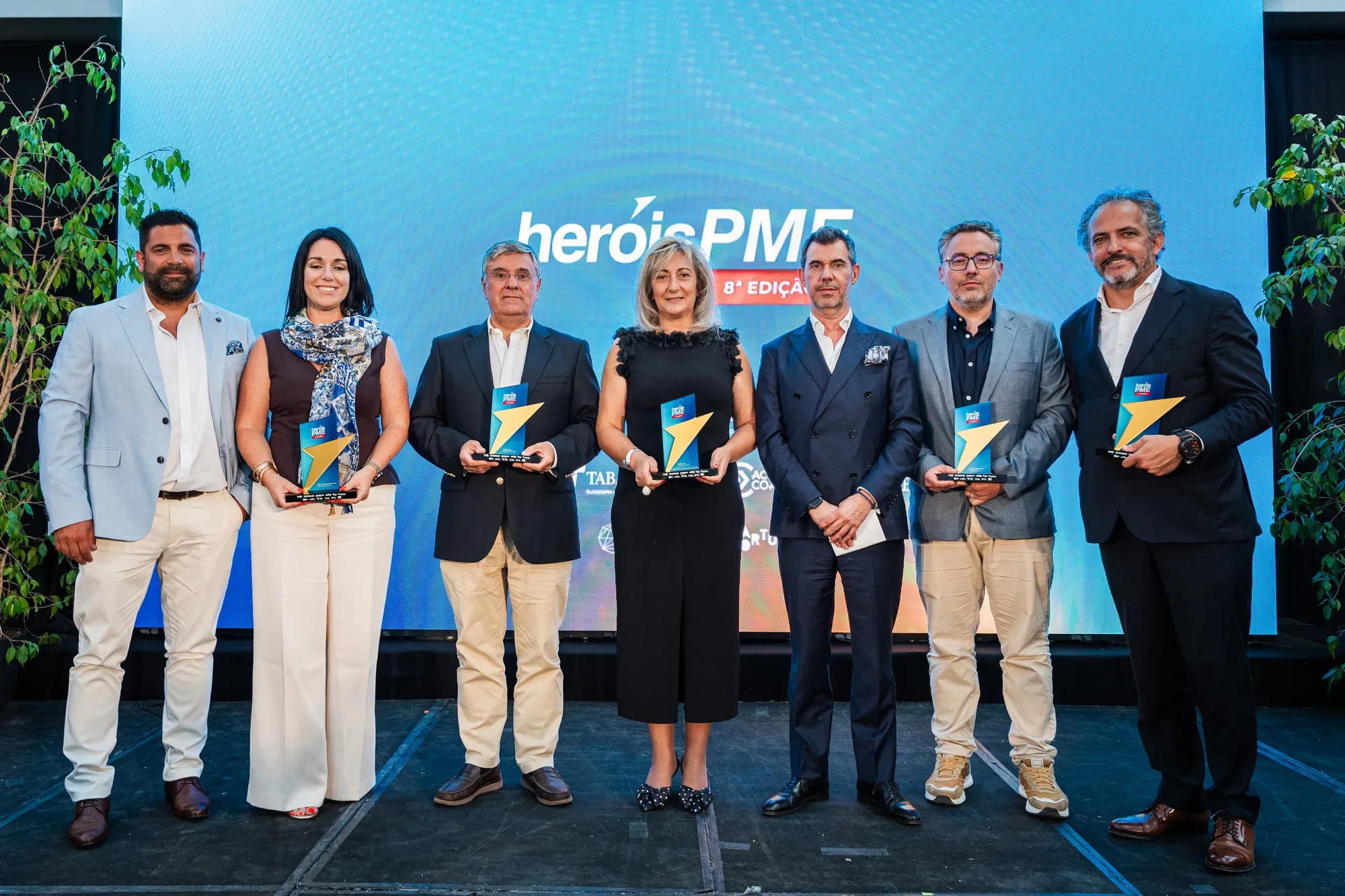 Grupo de sete pessoas posando no palco da cerimónia Heróis PME 8.ª Edição, alguns segurando troféus do prémio