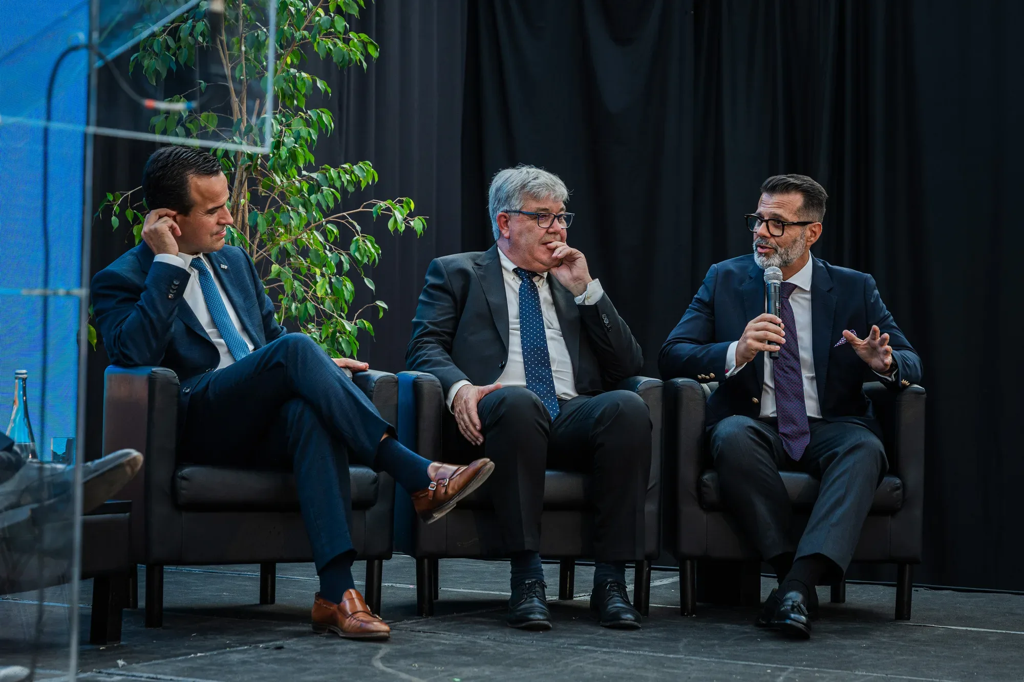 Três homens de fato sentados num painel de debate, com um deles a falar ao microfone enquanto os outros dois ouvem atentamente
