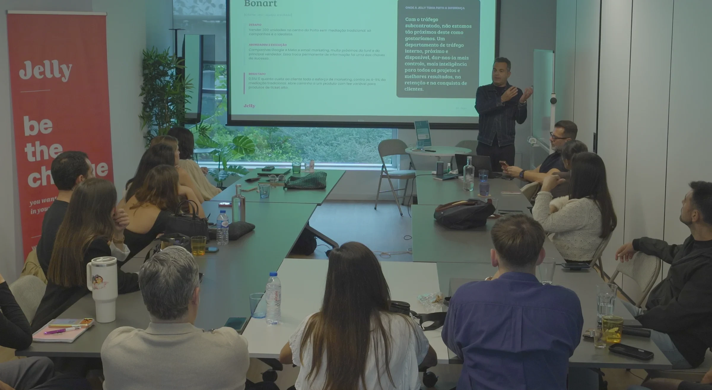 Homem apresenta slide "Bonart" a equipa sentada em mesa de reunião, com roll-up "Jelly — be the change" ao lado
