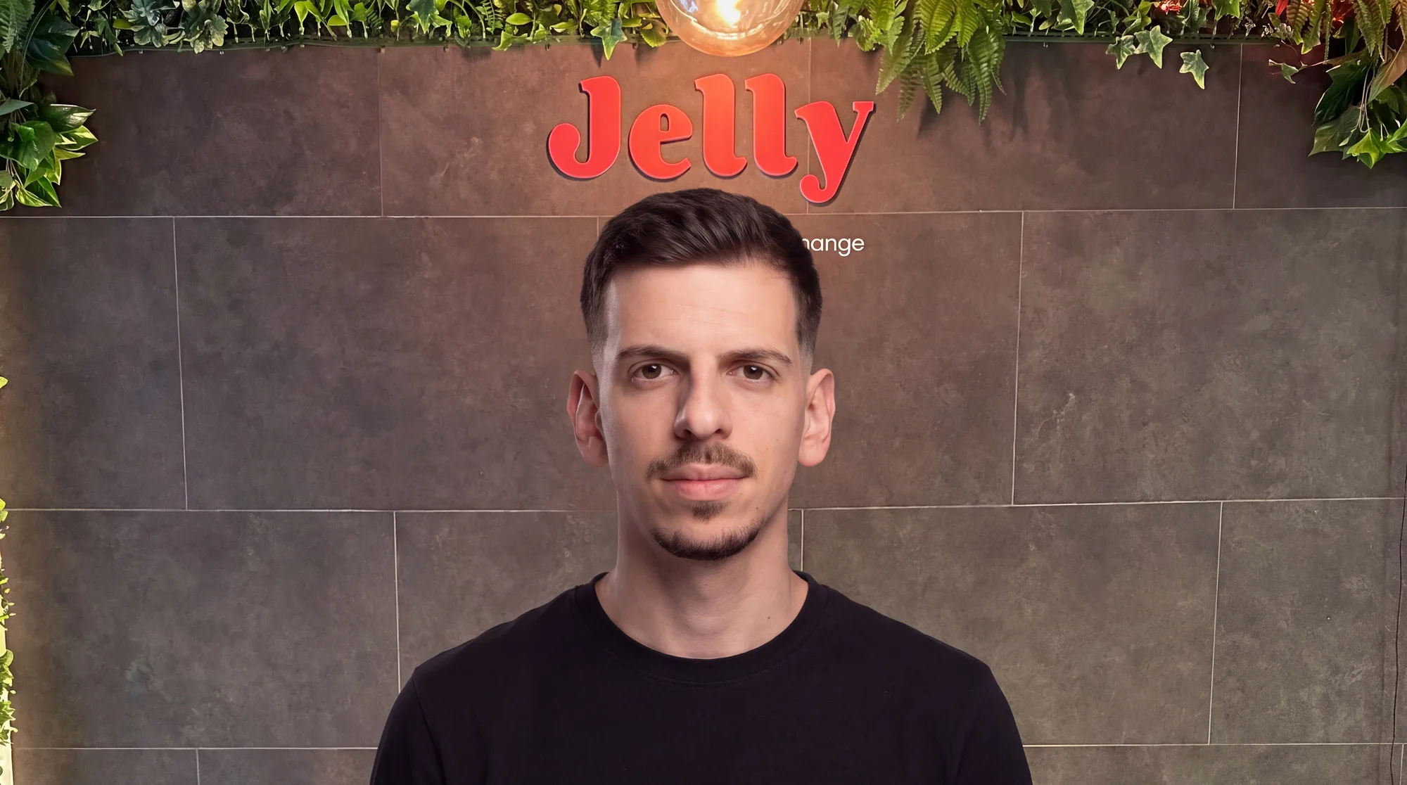 Homem de t-shirt preta diante de parede com o logótipo "Jelly" e plantas suspensas no topo